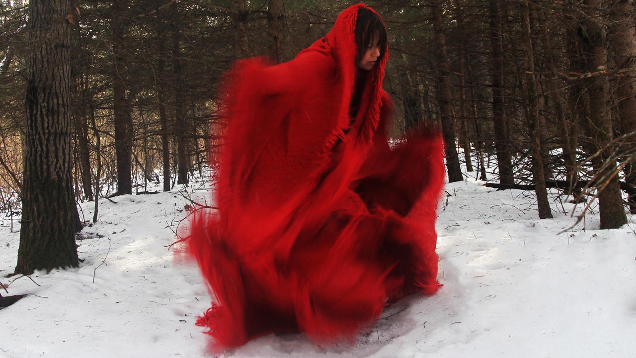 A photo shows a woman in movement, wearing a red blanket-like dress and surrounded by trees with snow on the ground.