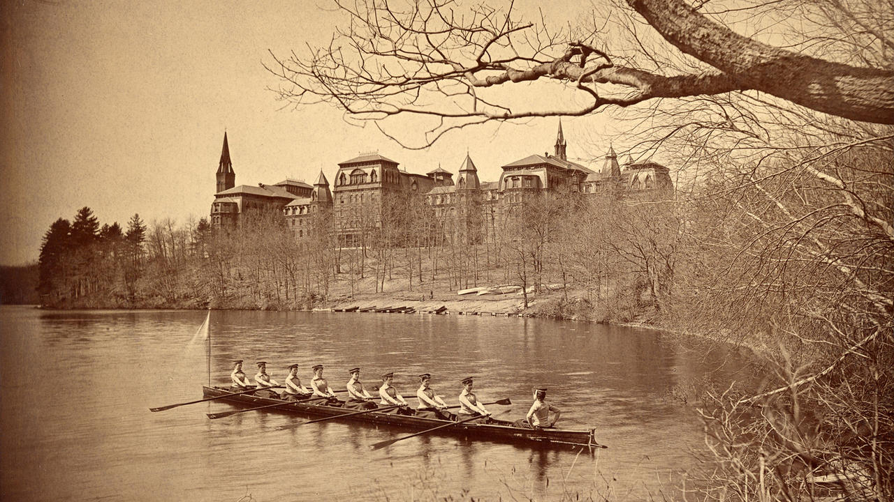 The class crew of 1894 rows on Lake Waban, with College Hall in the background.