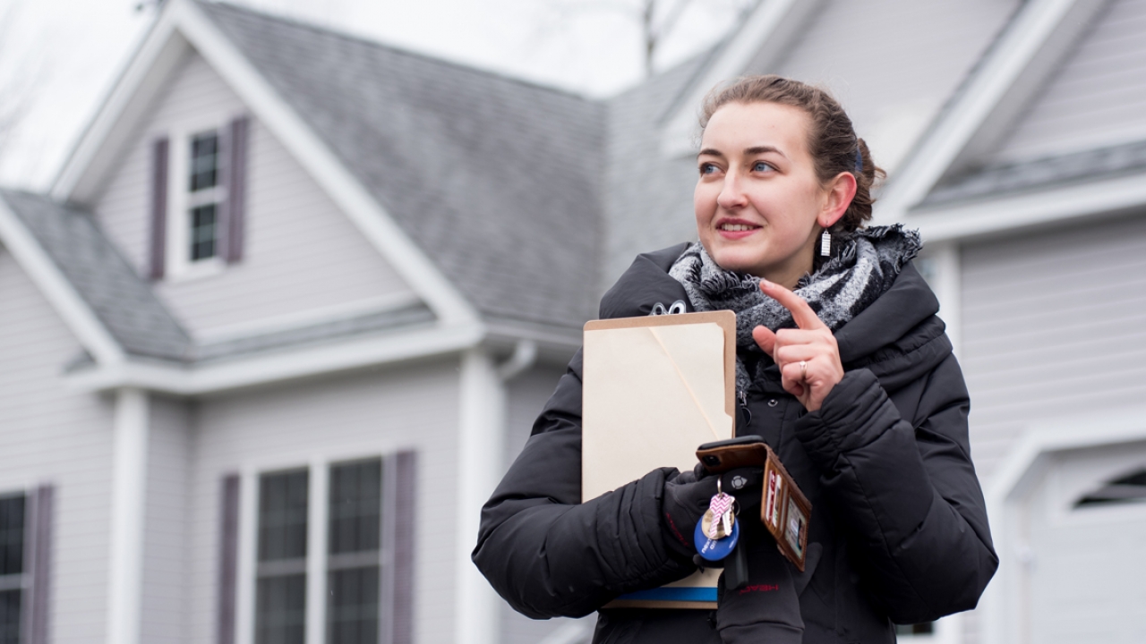 An alumna stands in front of a gray house with a clipboard