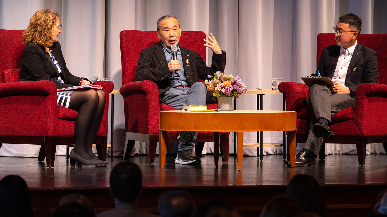 Haruki Murakami seated on stage and holding a microphone to answer a question.