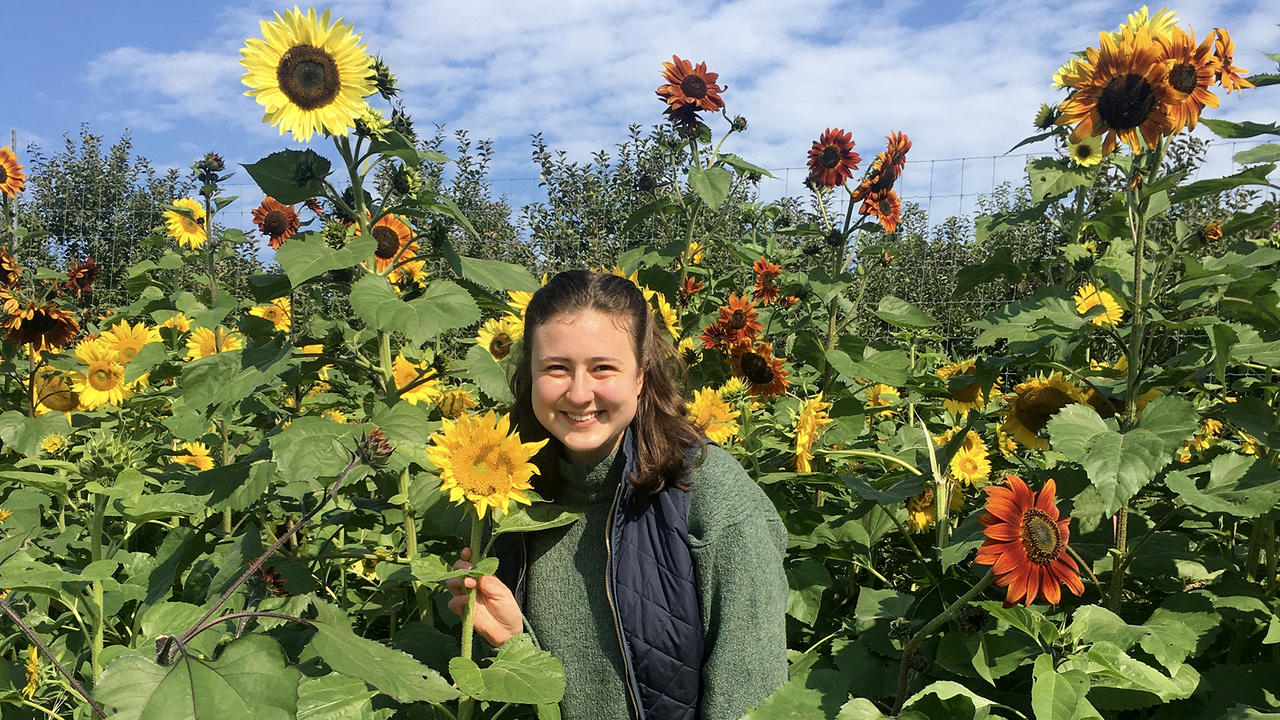 Dominique Mickiewicz stands in a garden of sunflowers and smiles at the camera.