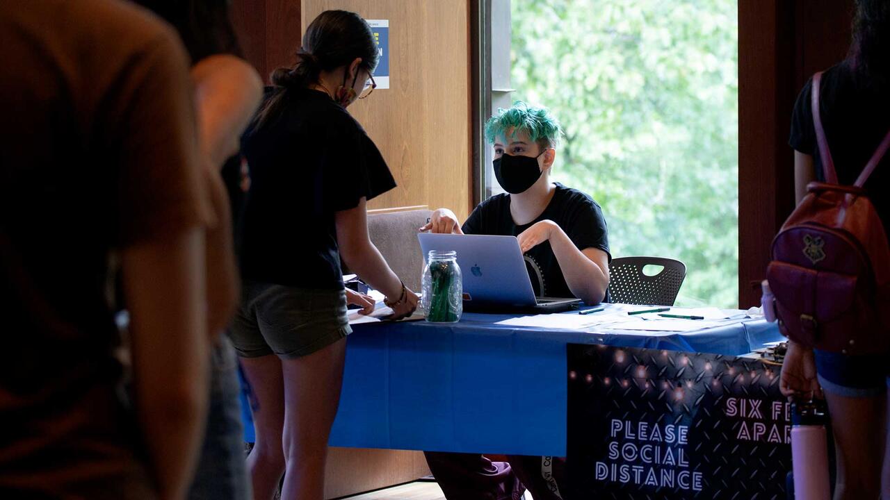 a person sitting behind a laptop wearing a mask speaking to another person across the table