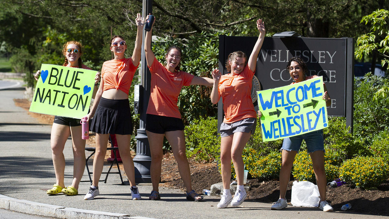 Five students stand at the entrance of Wellesley, waving welcome signs.