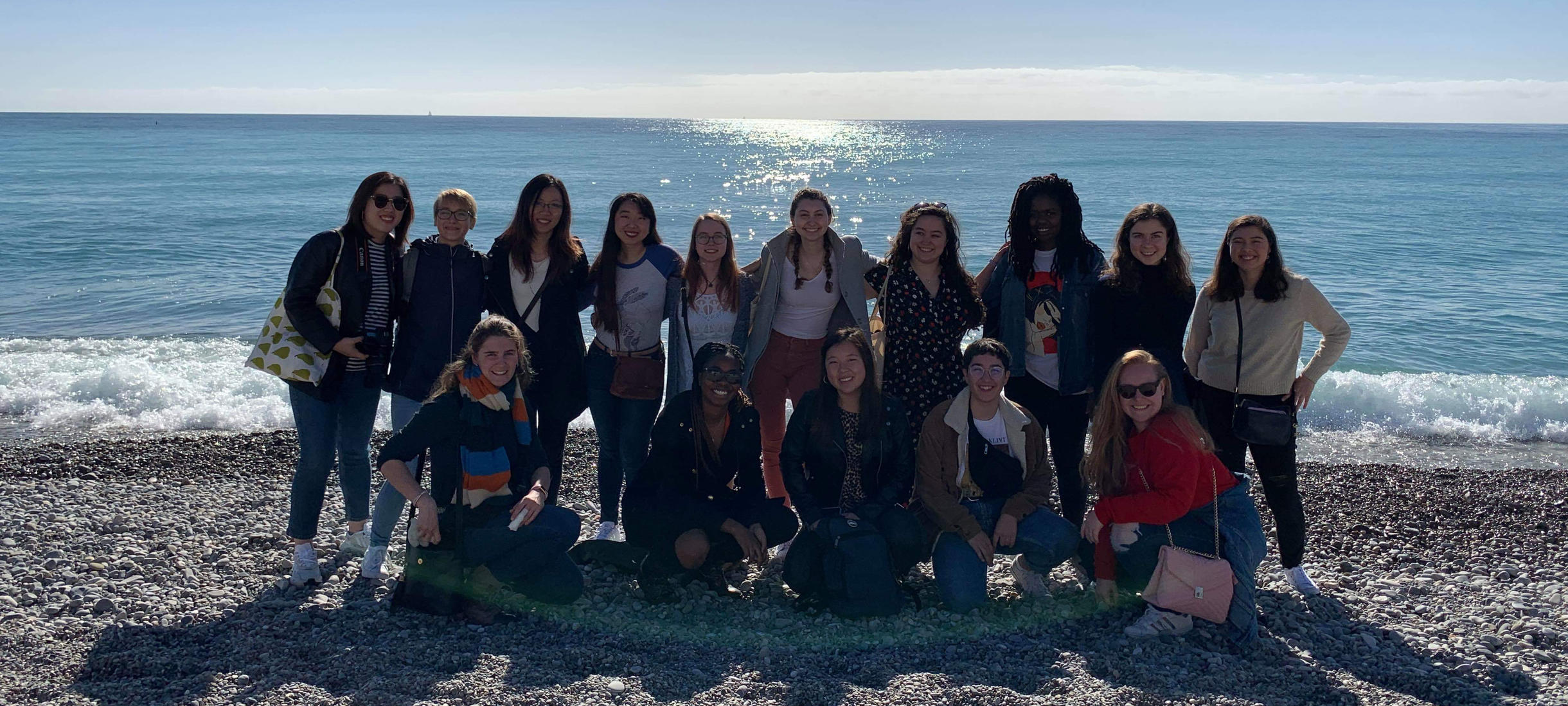 A smiling group of Wellesley-in-Aix students on a beach with water behind them, facing the sun.