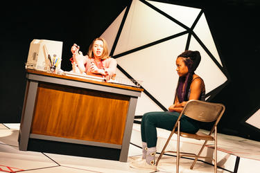Two actors at a desk and chair performing on stage.
