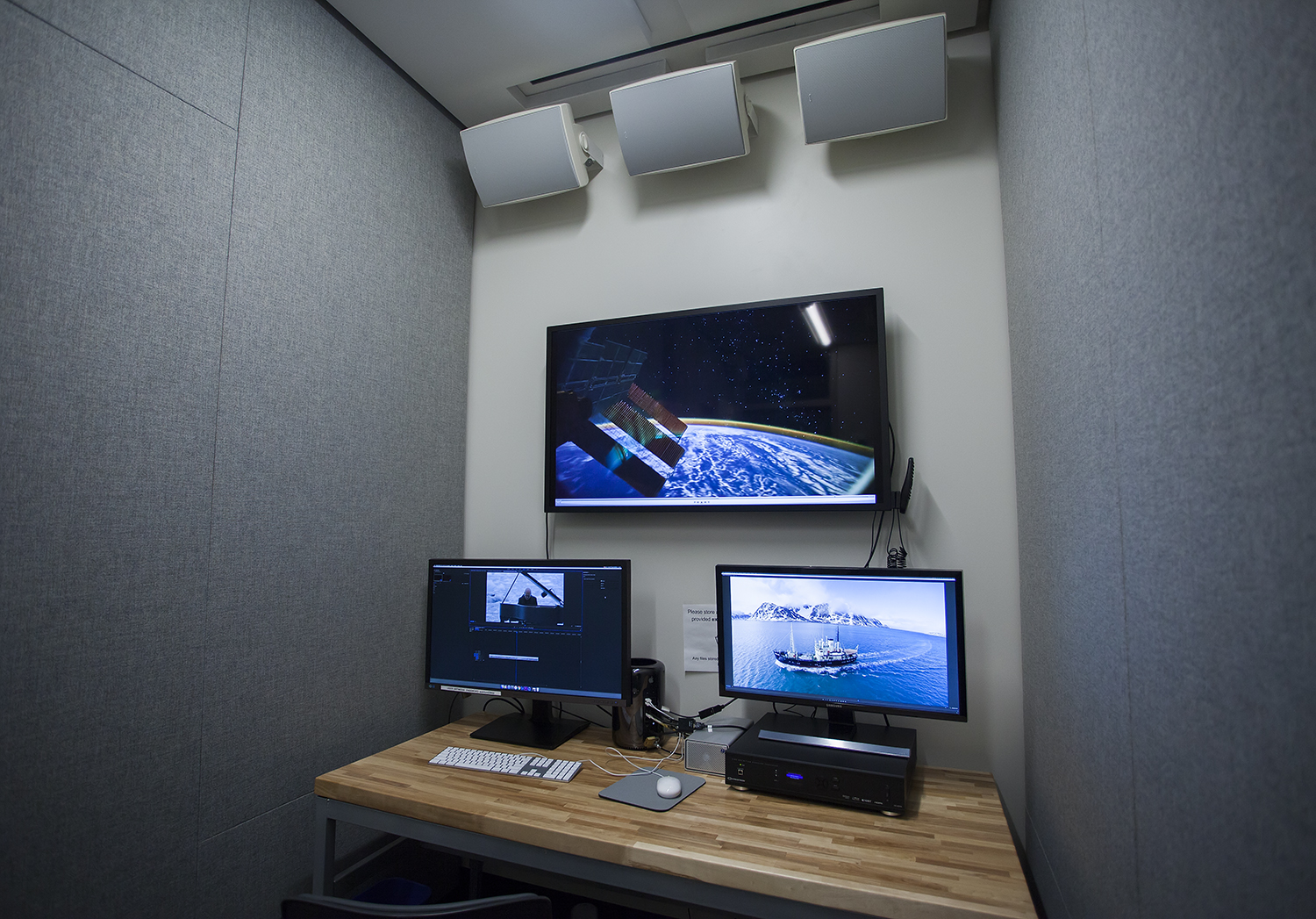 two computer monitors on a desk with one larger monitor on the wall behind them, three speakers mounted on the wall above that